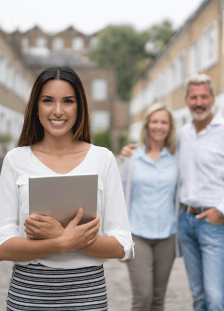 Agente inmobiliaria con una tablet en primer plano y una pareja sonriente de fondo frente a viviendas en hilera.